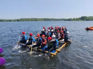 Large group of people in life jackets and helmets paddling a long raft on a lake, with blue water and green shoreline in the background.