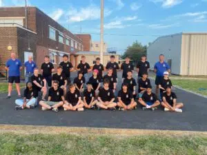 Group of students and a few adults posing for a photo outside a brick school building, all wearing matching black shirts with a small emblem on the left chest under a blue sky with clouds.