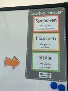 Laut-o-meter poster showing three colored blocks: Sprechen (normal voice), Flüstern (whisper), Stille (silence) with descriptions and an orange arrow on the left side.
