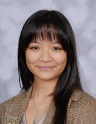 Portrait of a smiling Asian woman with long black hair, wearing a brown blazer against a gray studio backdrop.