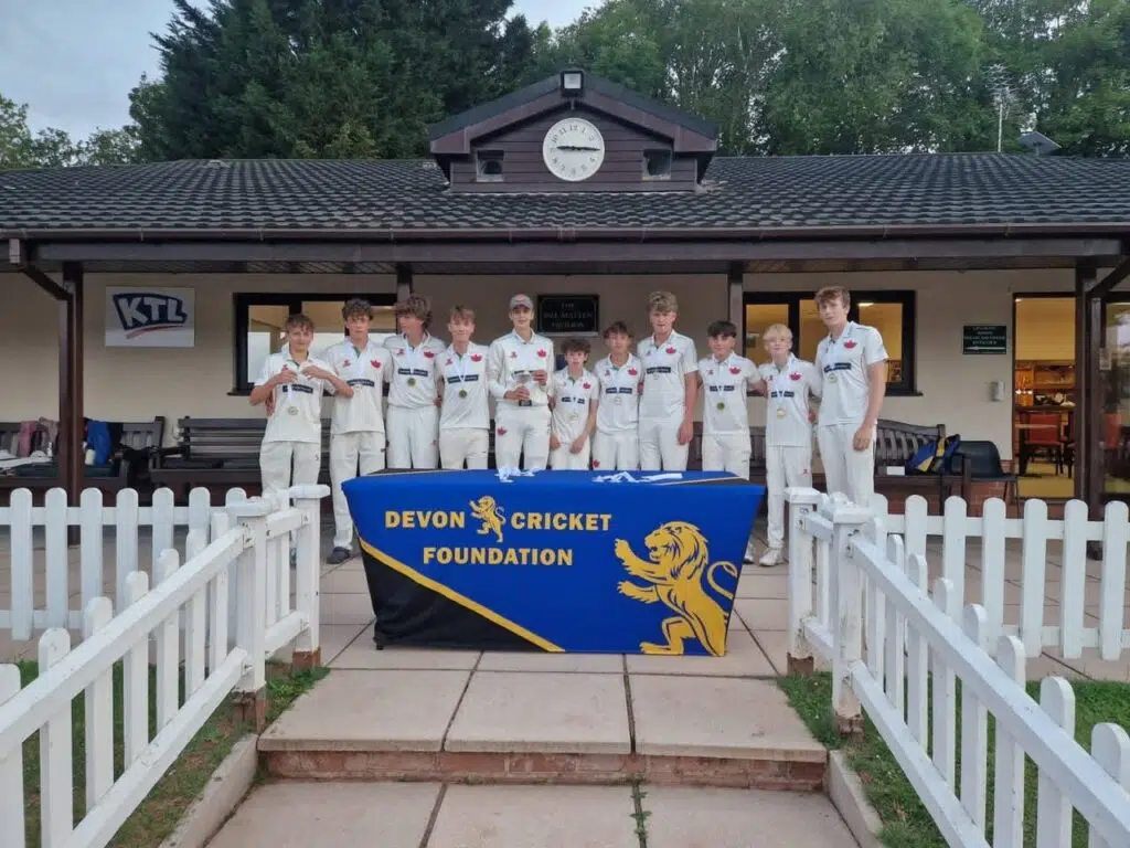 Group of young cricketers in white uniforms stand behind a table with a blue Devon Cricket Foundation banner and medals around their necks, outside a clubhouse with a clock above.