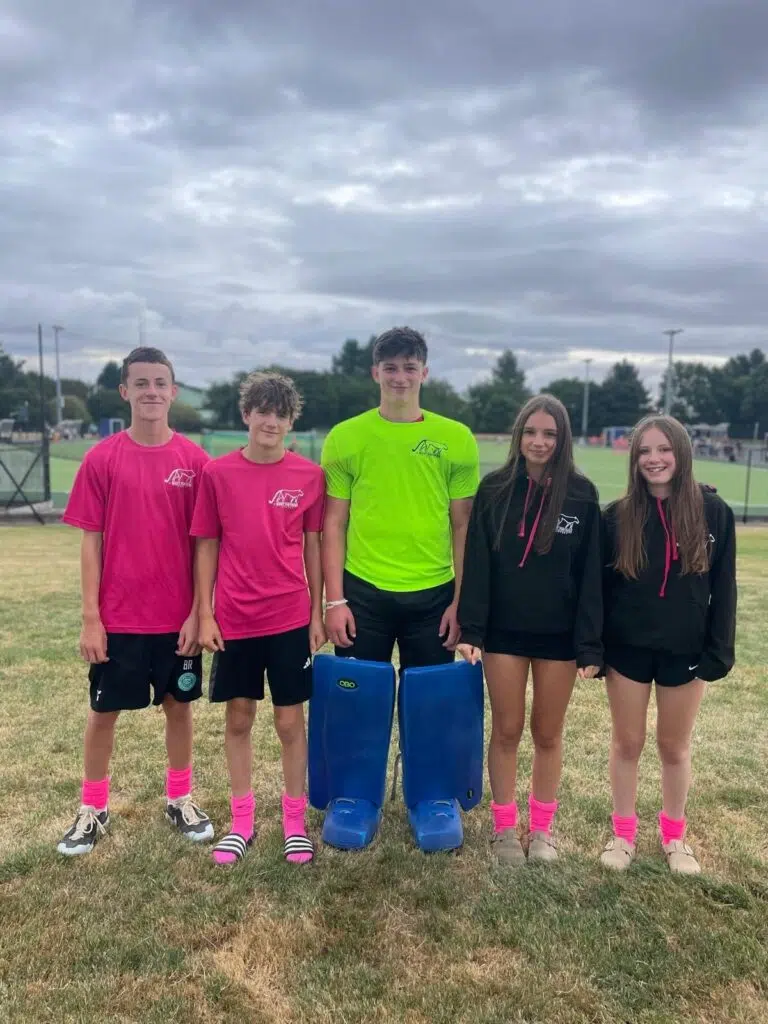 Five teens posing on a grassy sports field before a game: two in pink shirts, one in a neon lime shirt with blue goalie leg pads, and two girls in black hoodies.