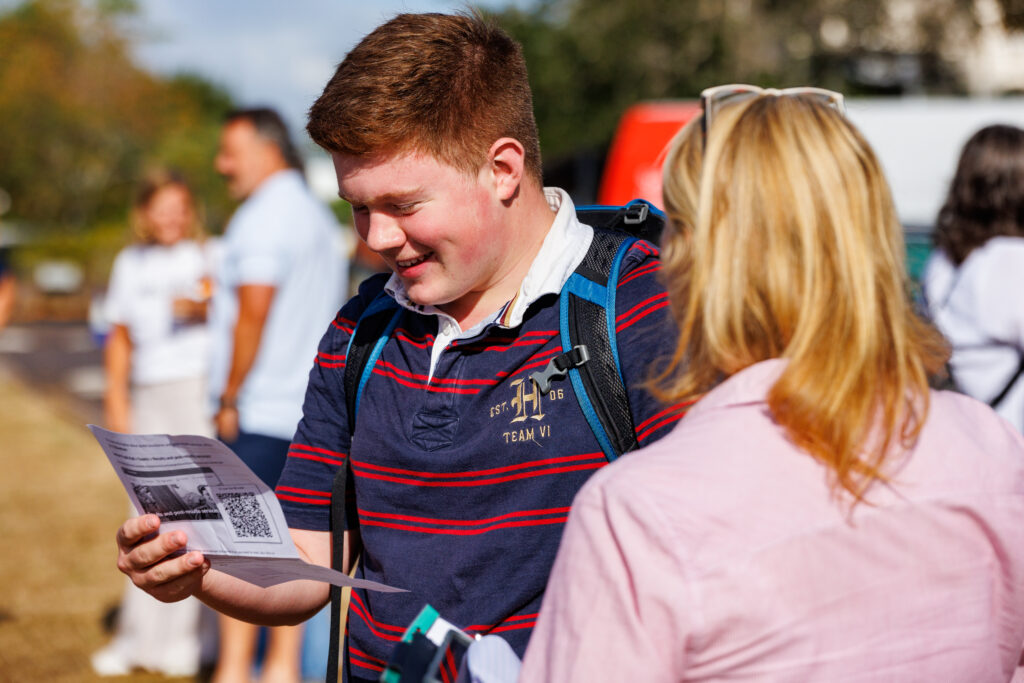 Young man with a backpack reads a flyer outdoors, smiling as others stand nearby.