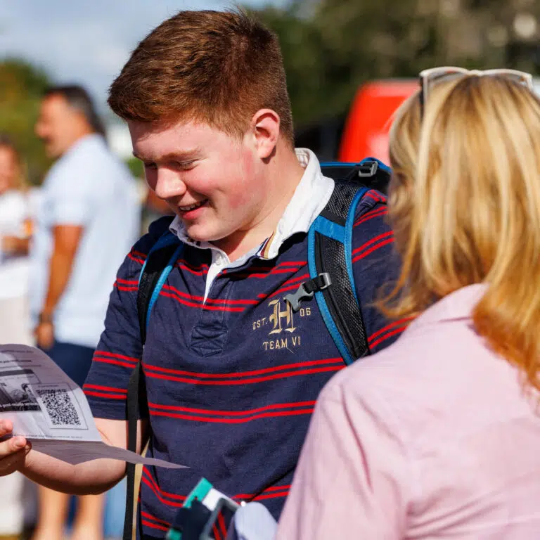 Young man with a backpack reads a flyer outdoors, smiling as others stand nearby.