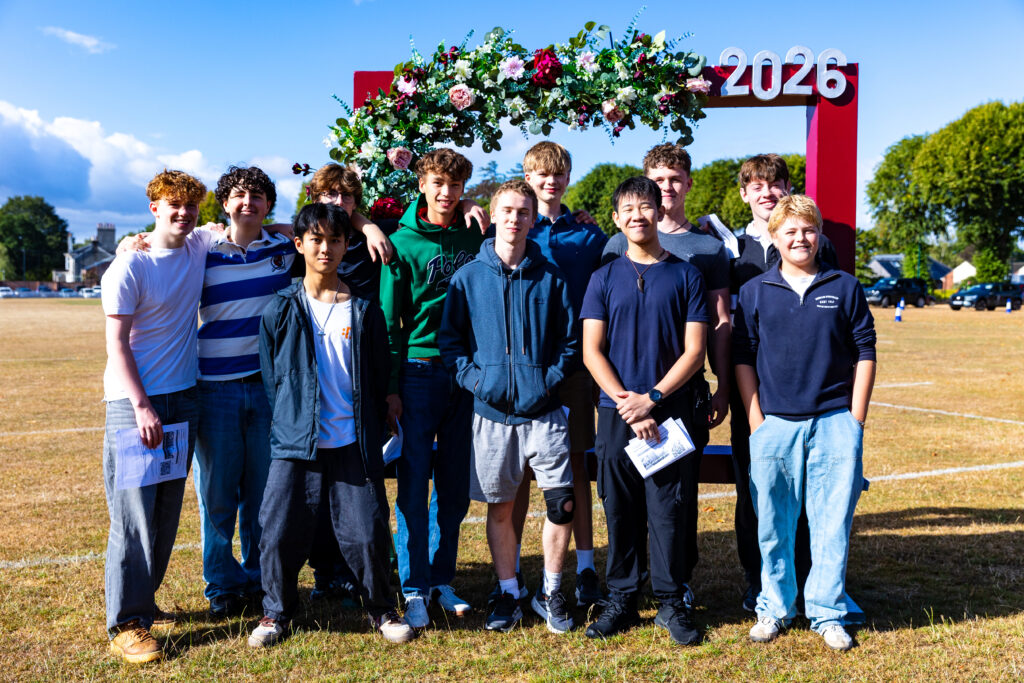 Group of teenagers posing together outdoors under a flower-covered arch with '2026' at the top on a sunny day.