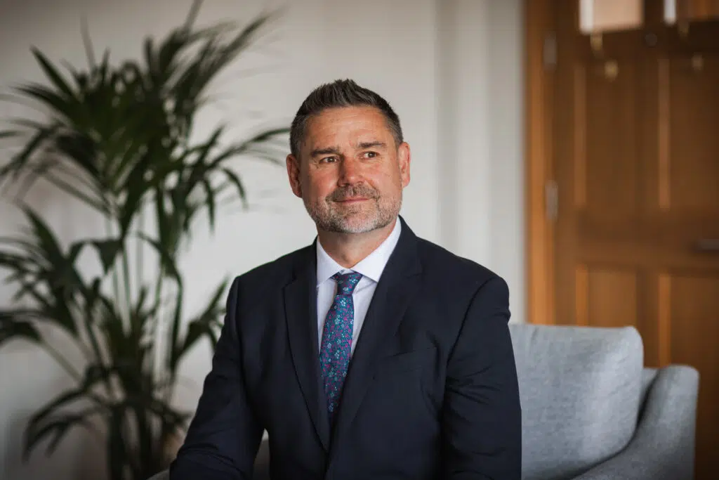 Professional man in a dark suit and patterned tie sitting on a couch in a modern office living area, looking thoughtfully to the side.