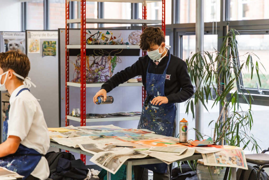 Student in a paint-splattered apron uses a spray can at a cluttered art table while wearing a mask in a bright classroom.