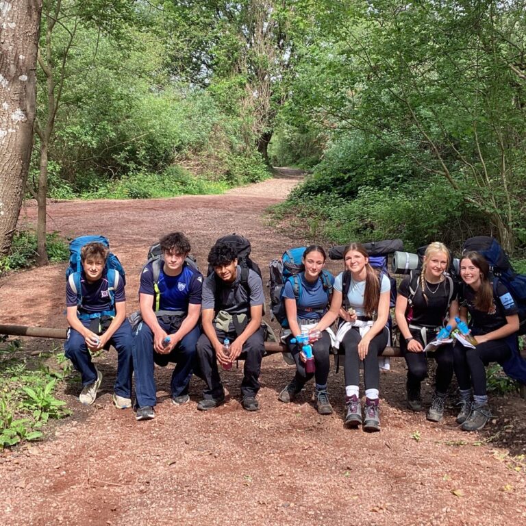 Group of young hikers with backpacks sitting on a log along a dirt forest trail, surrounded by trees and greenery.
