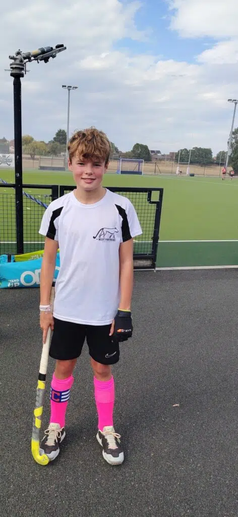 Young boy in a white sports shirt and black shorts stands on a field hockey pitch, holding a hockey stick and wearing bright pink socks and gloves.