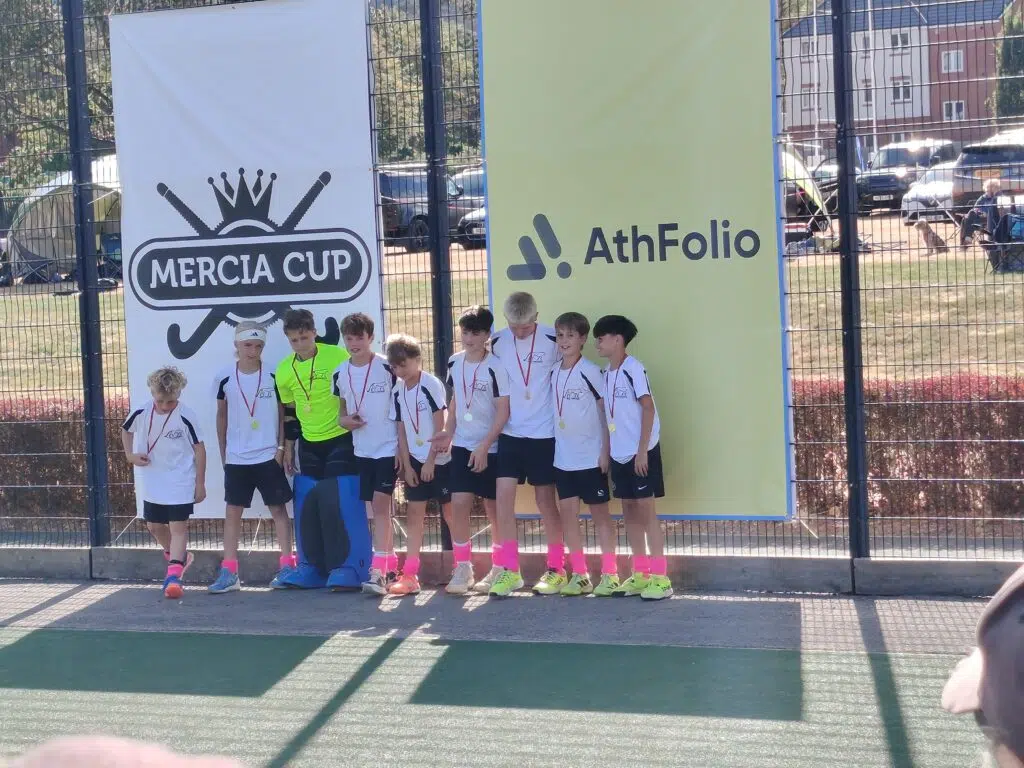 Young soccer team posing for a group photo in front of banners reading Mercia Cup and AthFolio, medals around their necks on a sunny day behind a fence.
