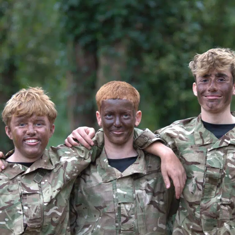 Three young people in camouflage uniforms with arms around each other, smiling outdoors in green, wooded background.