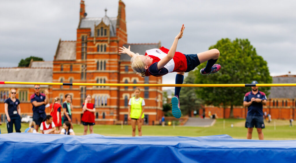Sports day high-jump activity set up on blue mats with yellow pole in front of big red brick school building with pupils jumping with bodies horizontal to camera.