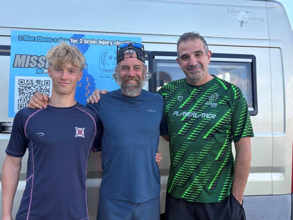 Three men in athletic shirts posing together in front of a van with a blue banner promoting a brain injury charity event.
