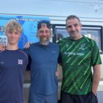 Three men in athletic shirts posing together in front of a van with a blue banner promoting a brain injury charity event.