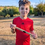 a bow in a red sports top holding drum sticks with grass and tress behind him