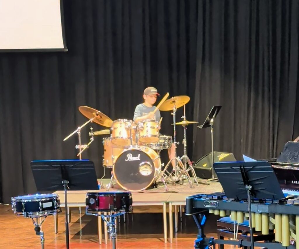 a young bow playing the drums on a stage with a black curtain behind him