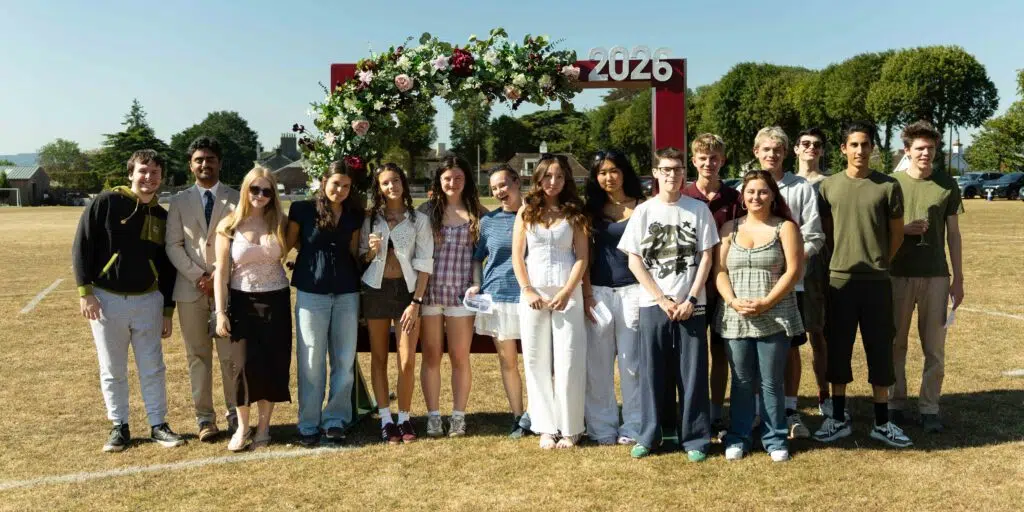 Group of teens posing for a class of 2026 photo under a flowered arch on a sunny sports field, about 20 people smiling at the camera
