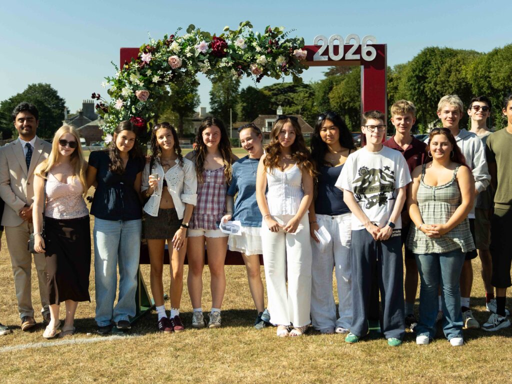 Group of teens posing for a class of 2026 photo under a flowered arch on a sunny sports field, about 20 people smiling at the camera