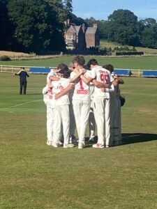 Cricket players in white uniforms form a huddle on a sunny field, with a large brick house in the background.