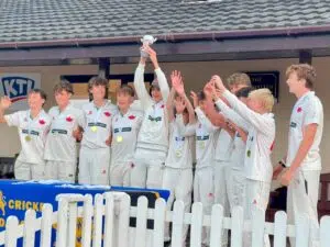 Junior cricket team celebrates victory, lifting a trophy aloft with gold medals around their necks in white uniforms with red logos to the side of a blue banner and white fence.