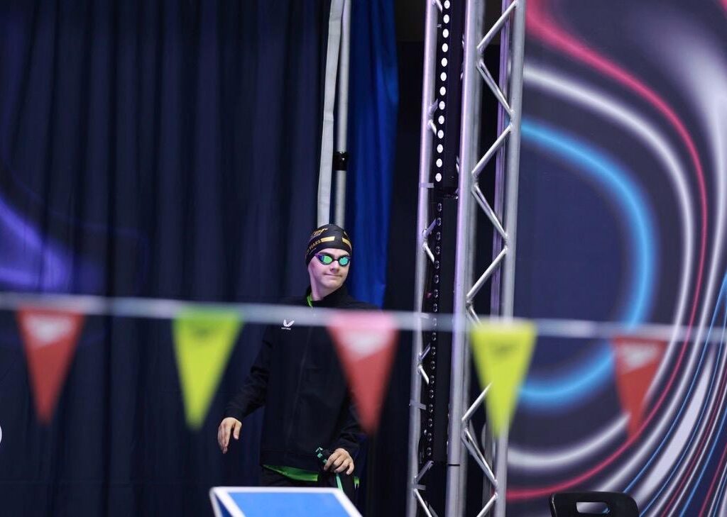 Swimmer wearing a cap and goggles stands backstage behind colorful pennant banners at an indoor swimming venue.