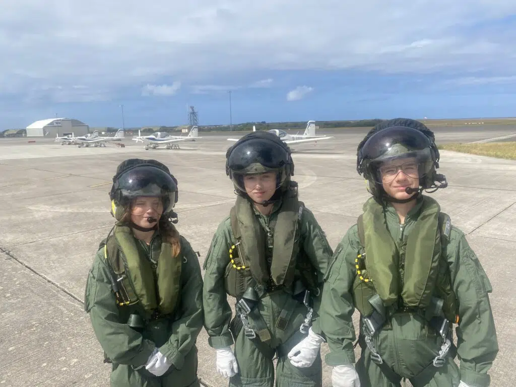 Three pilots in green flight suits and helmets stand at a runway, with small planes and hangars in the background.