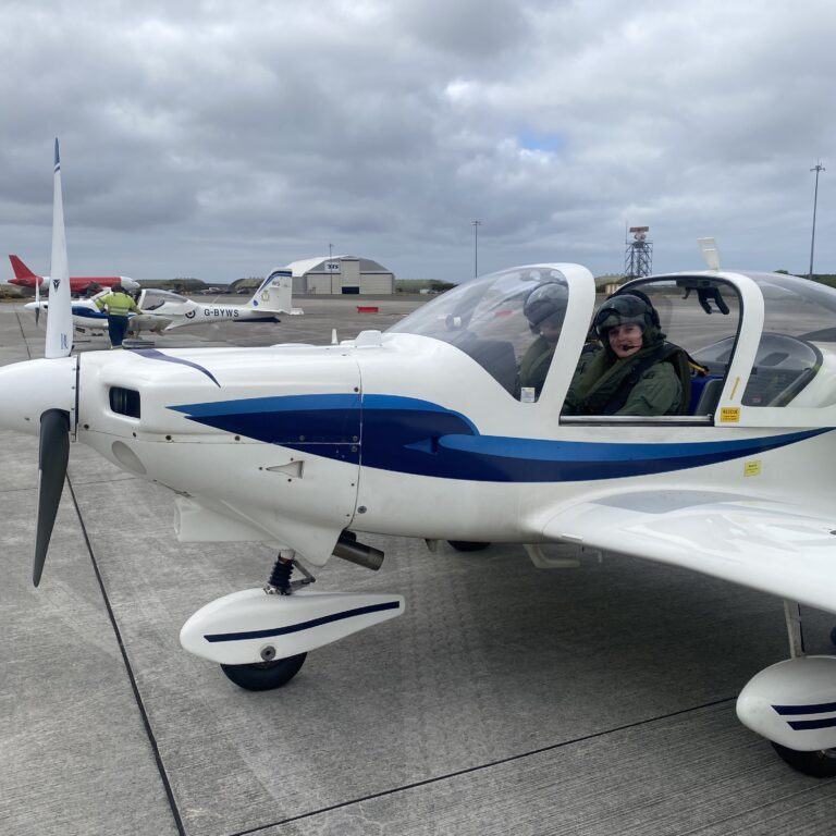 Small white and blue light aircraft on an airport tarmac with a pilot in the cockpit, foreground propeller visible.
