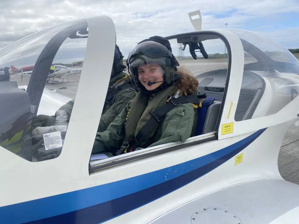 Smiling pilot wearing a helmet sits in the cockpit of a small white-and-blue training airplane on a runway.