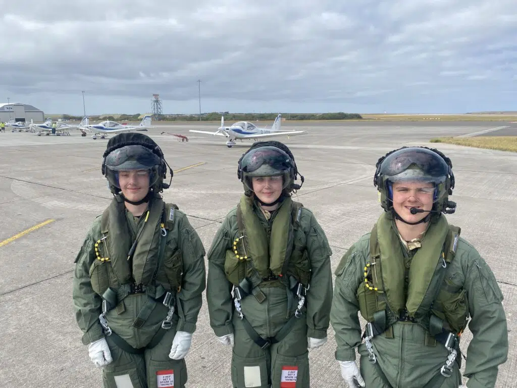 Three pilots in green flight suits and helmets stand on a concrete airfield with small white training aircraft in the background under a cloudy sky.”,