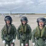 Three pilots in green flight suits and helmets stand on a concrete airfield with small white training aircraft in the background under a cloudy sky.”,